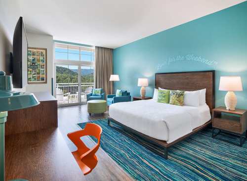 Bright hotel room with a king bed, colorful decor, and large windows showcasing mountain views.