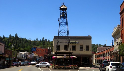 A small town street scene featuring a tall tower, shops, and green hills under a clear blue sky.