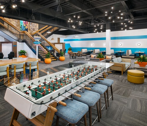 A modern lounge area featuring a foosball table, seating, plants, and a staircase with decorative lighting.