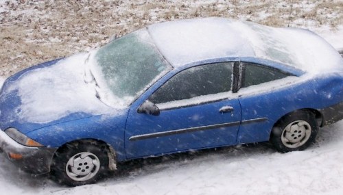 A blue car covered in snow, parked on a snowy street during a winter storm.