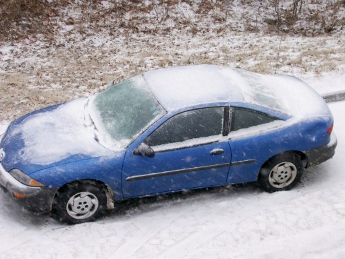 A blue car covered in snow, parked on a snowy road with trees in the background.