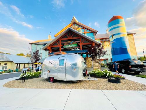 A shiny silver Airstream trailer in front of a colorful lodge with a large silo, surrounded by flowers and a parking area.