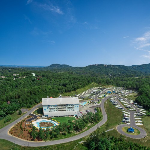 Aerial view of a hotel surrounded by greenery and a winding road, with parking and outdoor spaces visible.