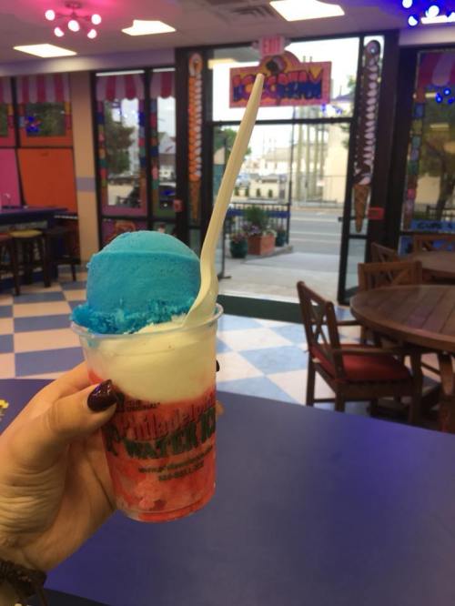 A hand holds a cup of colorful ice cream with blue and red layers, set in a vibrant ice cream shop.