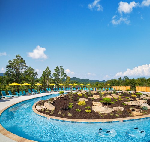A scenic pool area with lounge chairs, umbrellas, and landscaped rocks under a clear blue sky.