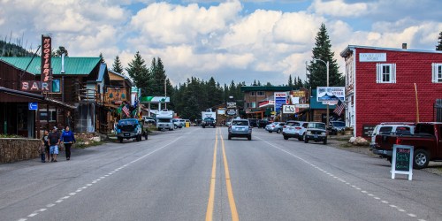 A small town street lined with shops and cars, surrounded by trees under a cloudy sky.