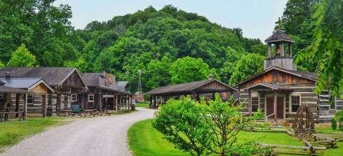 A rustic village with wooden buildings and a gravel path, surrounded by lush green trees and hills.