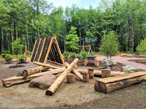 A natural playground featuring wooden structures, logs, and rocks, surrounded by trees and greenery.