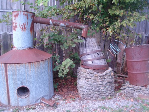 An old, rusty distillation apparatus made of metal and wood, surrounded by greenery and a stone base.