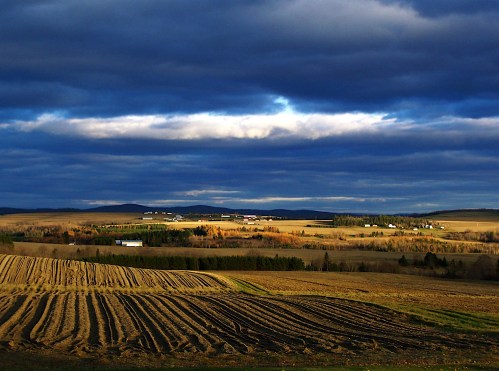 A scenic landscape featuring rolling fields, dark clouds, and distant hills under a dramatic sky.