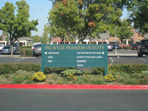 Sign at Vacaville Premium Outlets listing stores: Ann Taylor, Bass, Nike, Burberry, Coach, and bus tour drop-off location.