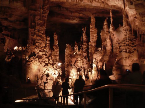 Dimly lit cave interior with towering stalagmites and stalactites, featuring silhouettes of visitors exploring the space.