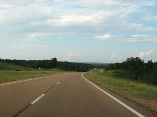A winding road stretches through green hills under a partly cloudy sky.