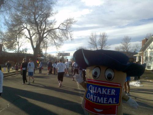 A mascot dressed as a Quaker Oats box stands on a street with people participating in an outdoor event.