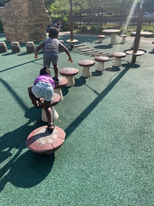 Two children play on a colorful playground, climbing on mushroom-shaped stepping stones. Sunlight casts long shadows.