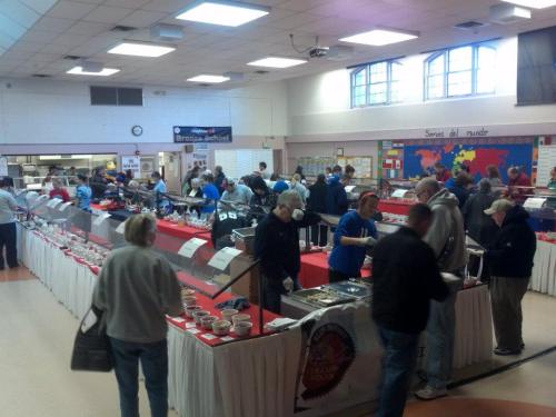 A busy community event with people browsing food displays and serving stations in a large hall.
