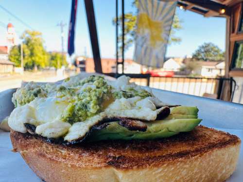 A toasted slice of bread topped with avocado, eggs, and green sauce, with a sunny outdoor setting in the background.