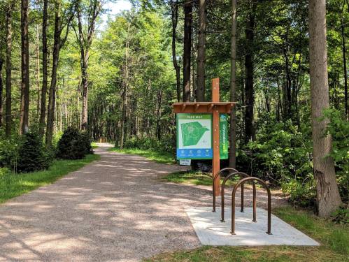 A park entrance with a map sign and bike rack, surrounded by lush green trees and a gravel path.
