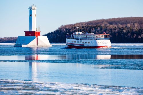 A boat navigates icy waters near a red and white lighthouse on a clear day, surrounded by wooded hills.