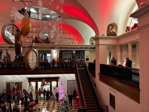 A festive indoor scene with holiday decorations, a large clock, and people gathered around a Christmas tree.
