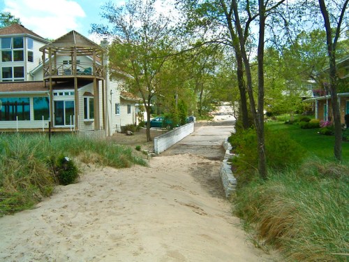 A sandy path leads between houses, with greenery and a wooden structure visible in the background under a blue sky.