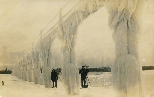 Historic photo of a frozen bridge, with people and a dog standing on snow, surrounded by icy structures.