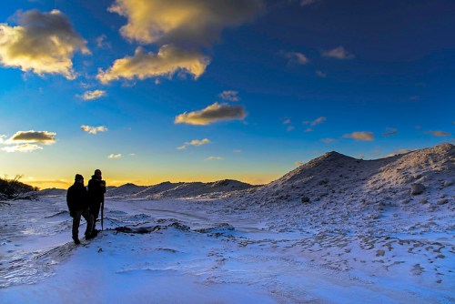 Two hikers stand on a snowy landscape, silhouetted against a vibrant sunset and dramatic clouds.