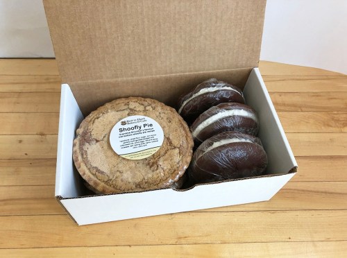 A white box containing a Shoofly pie and two wrapped chocolate desserts on a wooden surface.