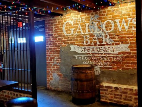 Interior of a bar with exposed brick walls, a wooden barrel, and string lights, featuring a vintage sign for "The Gallows Bar."