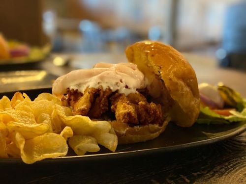 A close-up of a crispy chicken sandwich with creamy sauce, served with a side of potato chips on a black plate.