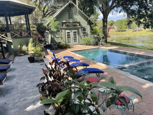 A serene backyard scene featuring a pool, colorful lounge chairs, and a charming green house by a lake.