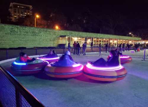 Colorful bumper cars spin on an ice rink at night, with spectators and lights in the background.