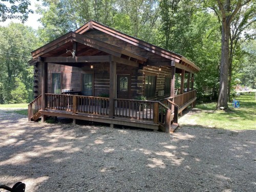 A rustic wooden cabin surrounded by trees, featuring a spacious porch and gravel pathway.