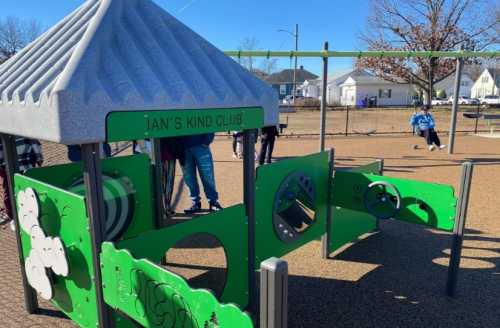 A colorful playground structure named "Ian's Kind Club" with green panels and a slide, set in a sunny park.