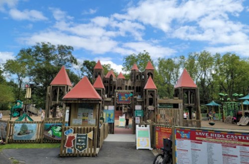 A large wooden playground with red-roofed towers, surrounded by trees and blue skies, featuring various play structures.