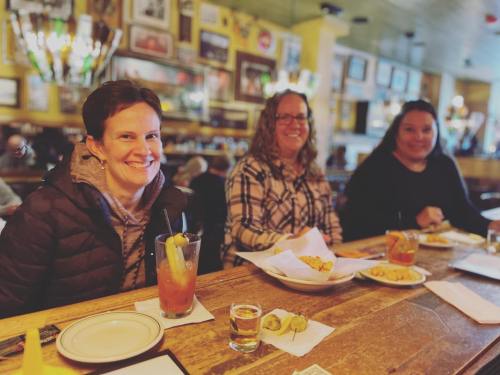 Three women sit at a bar, smiling, with drinks and food in front of them, surrounded by a cozy, decorated atmosphere.