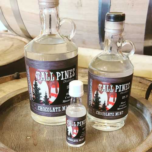 Three glass bottles of "Tall Pines" chocolate mint distillate on a wooden barrel, showcasing different sizes.