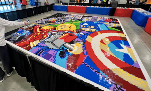 A large, colorful LEGO mosaic featuring superhero characters, displayed at an event with tables in the background.