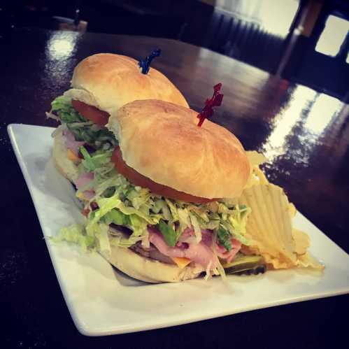 A plate with two stacked sandwiches topped with lettuce, tomato, and pickles, served with potato chips.