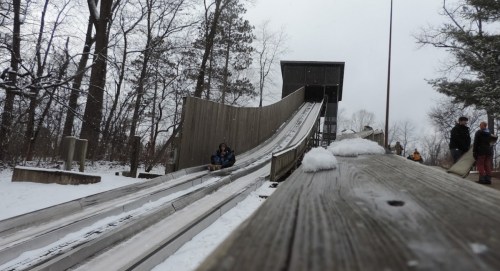 A snowy sledding hill with a wooden slide and people waiting at the top, surrounded by trees.