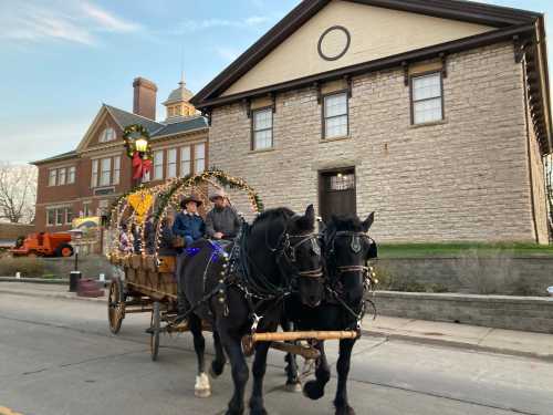 A festive horse-drawn wagon decorated with lights passes by a historic stone building. Two people ride in the wagon.