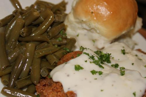 A plate featuring fried chicken smothered in white gravy, green beans, and a soft dinner roll.