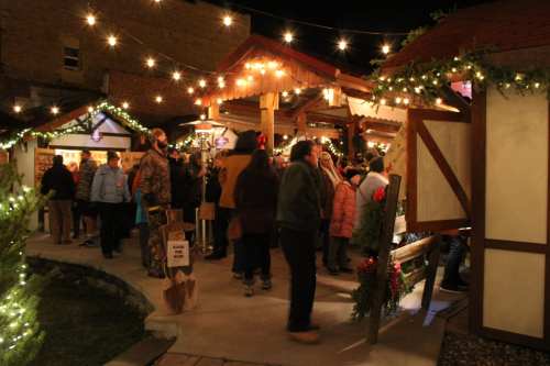 A festive outdoor market at night, with people browsing stalls decorated with lights and greenery.