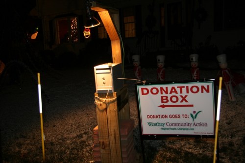 A wooden donation box with a sign for Westbay Community Action, illuminated at night with holiday decorations in the background.