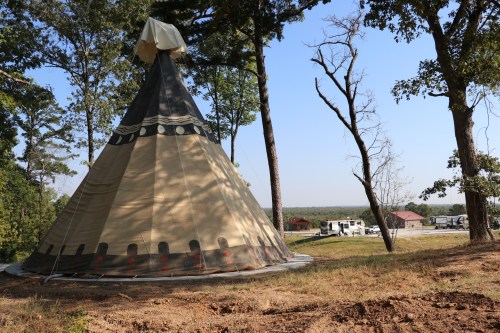 A large teepee stands in a grassy area surrounded by trees, with distant buildings and RVs visible in the background.