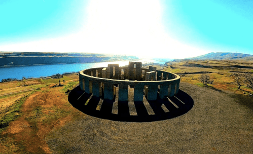 Aerial view of a stone circle structure near a river, surrounded by grassy fields and a bright blue sky.