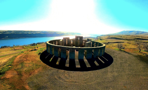 Aerial view of a stone circle structure near a river, surrounded by grassy fields and a bright blue sky.