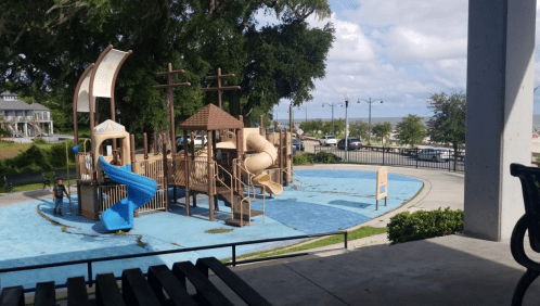 A playground with a water feature, slides, and climbing structures, surrounded by trees and a view of the street.