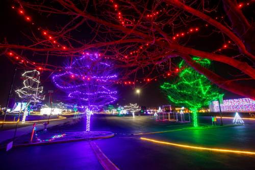 Colorful holiday lights illuminate trees and a parking lot at night, creating a festive atmosphere.
