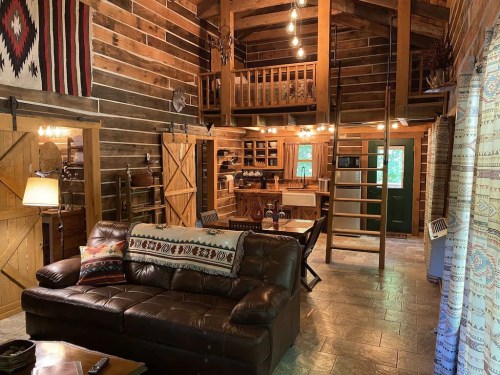 Cozy cabin interior with wooden walls, a leather sofa, and a lofted area, featuring warm lighting and rustic decor.